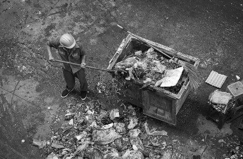 A worker wearing a white safety helmet and dark clothing is standing on a paved area in front of a large open-top industrial waste skip filled with mixed refuse, including paper, plastic, and other debris. The worker holds a long-handled tool, possibly a broom or rake, and is positioned to the left of the skip, which is situated on a rough, textured surface. In front of the worker, there are loose piles of rubbish scattered across the ground, consisting of crumpled paper, plastic bottles, and miscellaneous waste materials. The environment appears to be an outdoor space, likely a commercial or industrial site, with no greenery visible. The scene is in black and white, emphasizing the textures of the waste, the skip, and the surface. The image reflects a waste management or rubbish removal activity, which Garden Clearance Coney Hall offers as part of its garden clearance and rubbish disposal services in the local area.
