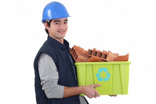 A young man with dark hair, wearing a blue safety helmet and a navy jacket over a grey long-sleeve shirt, is holding a bright green recycling bin with a blue recycling symbol. The bin contains a neatly stacked pile of red clay bricks with rectangular holes, which appear to be construction or garden materials. The man is smiling gently and is positioned against a plain white background. This image is relevant to rubbish removal services, such as those offered by Garden Clearance Coney Hall, which specialise in garden clearance and waste disposal in the local area near Coney Hall. The bricks and bin are clearly visible, with the bricks stacked inside the bin and the man's hand supporting the bottom. The overall scene suggests the collection or disposal of garden or building waste, suitable for inclusion on a website page about rubbish removal in the local postcode area, with a focus on environmentally responsible disposal methods.