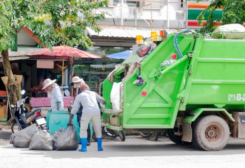 A green rubbish collection vehicle is parked alongside a busy street, with two sanitation workers dressed in light grey uniforms, blue rubber boots, and protective gloves loading black garbage bags into the vehicle. One worker is bending down to lift a bag, while the other stands nearby. The background includes a small shop with a red and white awning, potted plants, and several parked motorcycles. The scene is outdoors, with trees and urban infrastructure visible, indicating a waste removal operation in a town or suburbs near Coney Hall. The lighting suggests daytime with natural light illuminating the scene, highlighting the textures of the trash bags, the vehicle's metallic surface, and the surroundings. This image relates to rubbish collection services offered by Garden Clearance Coney Hall, focusing on waste removal in the local area.