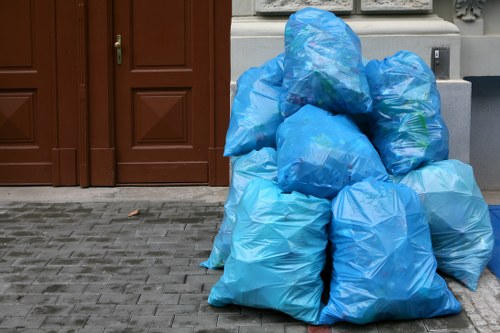 A group of large blue plastic rubbish bags filled with waste, stacked on a paved outdoor surface in front of a brown wooden door and a grey wall. The bags appear full and are made of lightweight material with a slightly glossy finish, some with visible creases and folds. The scene is set on a grey brick driveway, possibly part of a residential or commercial property, and the bags are arranged in a somewhat haphazard manner, indicating they are ready for collection or disposal. The background includes a closed wooden door with vertical panels and a small, off-white wall section with some decorative moldings, suggesting a tidy, well-maintained property in the local area. This image exemplifies rubbish collection activities carried out by Garden Clearance Coney Hall, a professional waste management service in the Coney Hall region.