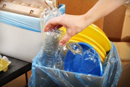 A close-up view of a person's hand holding a clear plastic water bottle with a ridged surface, positioned above a large blue rubbish bag that is partially open, revealing several yellow and blue plastic cleaning buckets stacked inside. The background shows part of a room with visible cardboard boxes and a black surface, indicating an indoor setting possibly used for storage or a rubbish collection area. The scene relates to waste disposal or rubbish removal, typical of services offered by companies like Garden Clearance Coney Hall in the local area. The plastic materials and buckets are standard household waste items, and the lighting appears natural, with no outdoor elements visible, emphasizing the focus on waste segregation or collection preparations.