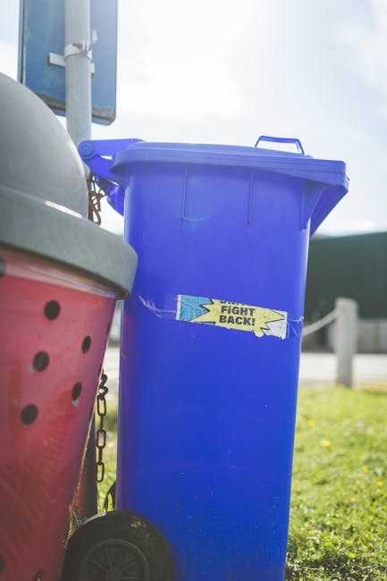 A bright blue wheeled rubbish bin with a closed lid is positioned outdoors on a paved surface, with a small yellow and blue sticker featuring the text 'FIGHT BACK!' partially torn and attached to its 