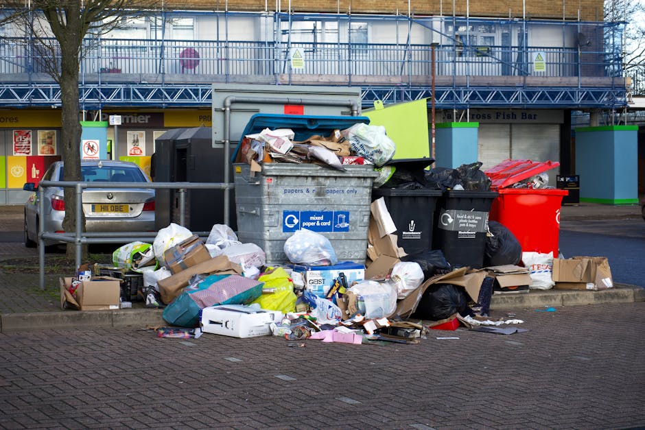 A bright blue wheeled rubbish bin with a closed lid is positioned outdoors on a paved surface, with a small yellow and blue sticker featuring the text 'FIGHT BACK!' partially torn and attached to its 